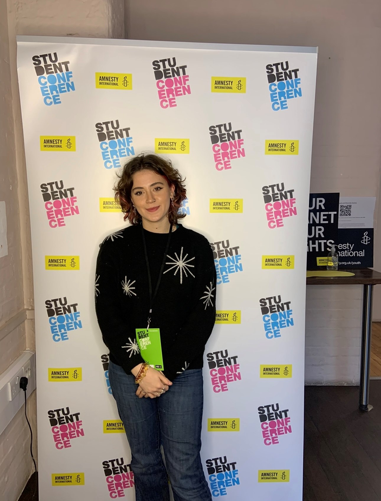 Picture of Kate Wilson. They are smiling, wearing a jumper with stars on it. Behind them is a roller banner that reads 'Amnesty International' and 'Student Conference'