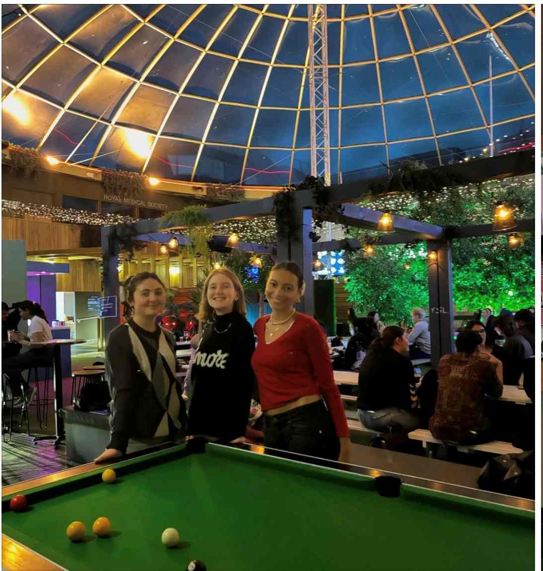 A photo of Mia Hiraani-Nelson with two frinds by a pool table in Potterow, the University of Edinburgh Student Union. Behind them is a dome and a lot of people socializing on benches.