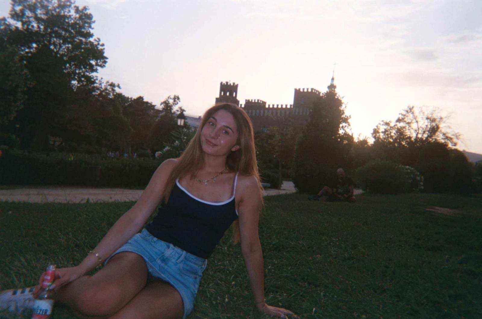 Photo of Mia Taylor sitting in a park. Behind her are some trees and a building that looks similar to a castle.