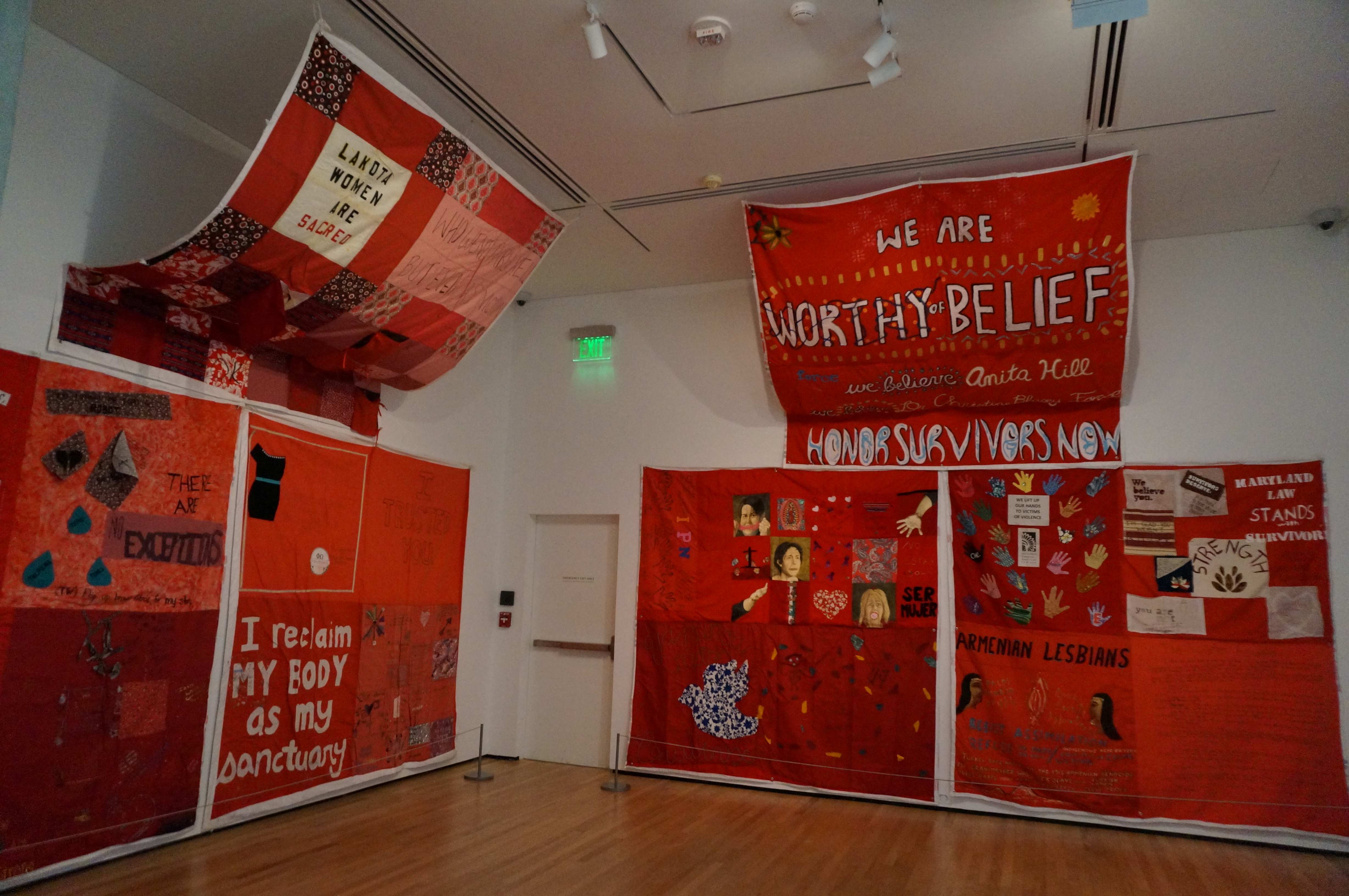 Photo of the monument quilt. It consists of multiple squares of fabric hanging on a wall or from the ceiling. Each square of The Monument Quilt carries a story of survival, solidarity, and healing.