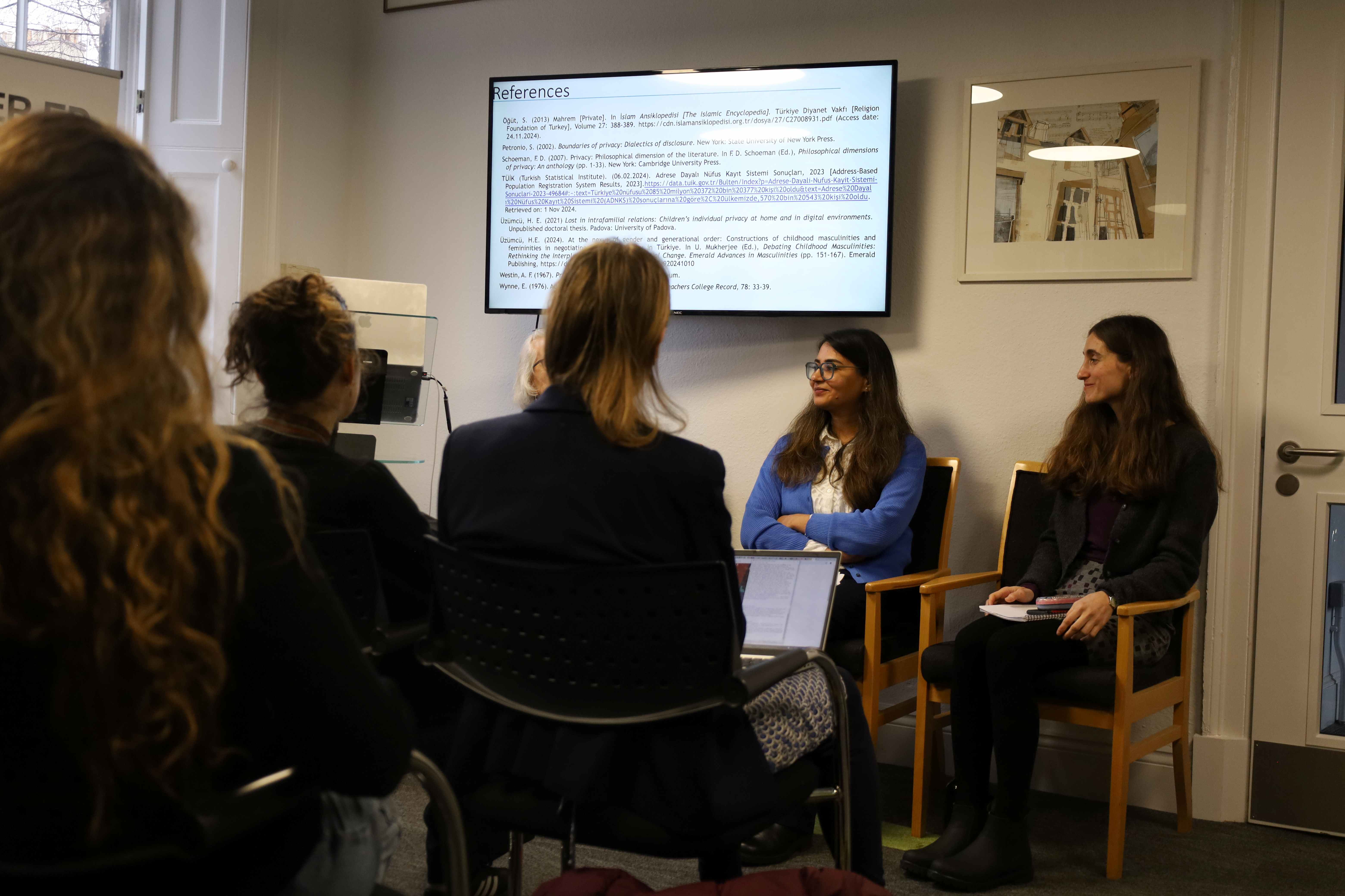 Photo of Sarah, Elif, and Patricia Jefferey at the International Women's Day event during the panel discussion. They are sitting in a row under a screen that reads 'references'. There are sitting in front of a group of an audience.