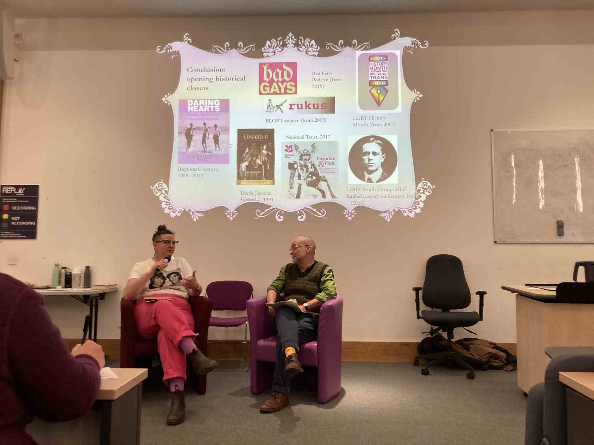 Photo of Professor Matt Cook and the panelists in Meadows Lecture Theatre sitting on two chairs, having a conversation. Behind them there is a screen with various images of podcasts, archives, and projects on 'opening the historical closet'.