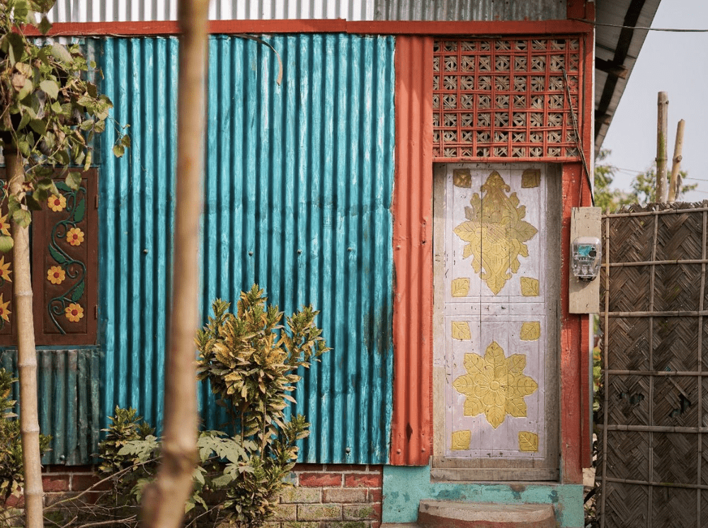 Image of a side of a home painted blue, with a pink and gold door. Trees and bushes overlap with the home in this image.