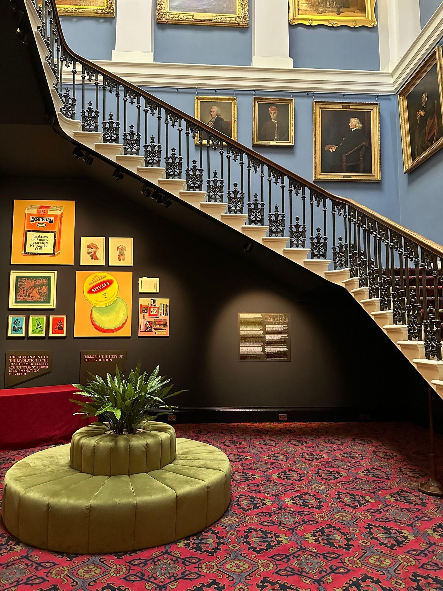Display of art works at the Old College of the University of Edinburgh. The display is located below a staircase and consists of a variety of vibrant and colourful pieces. There is a plant across the artwork. 