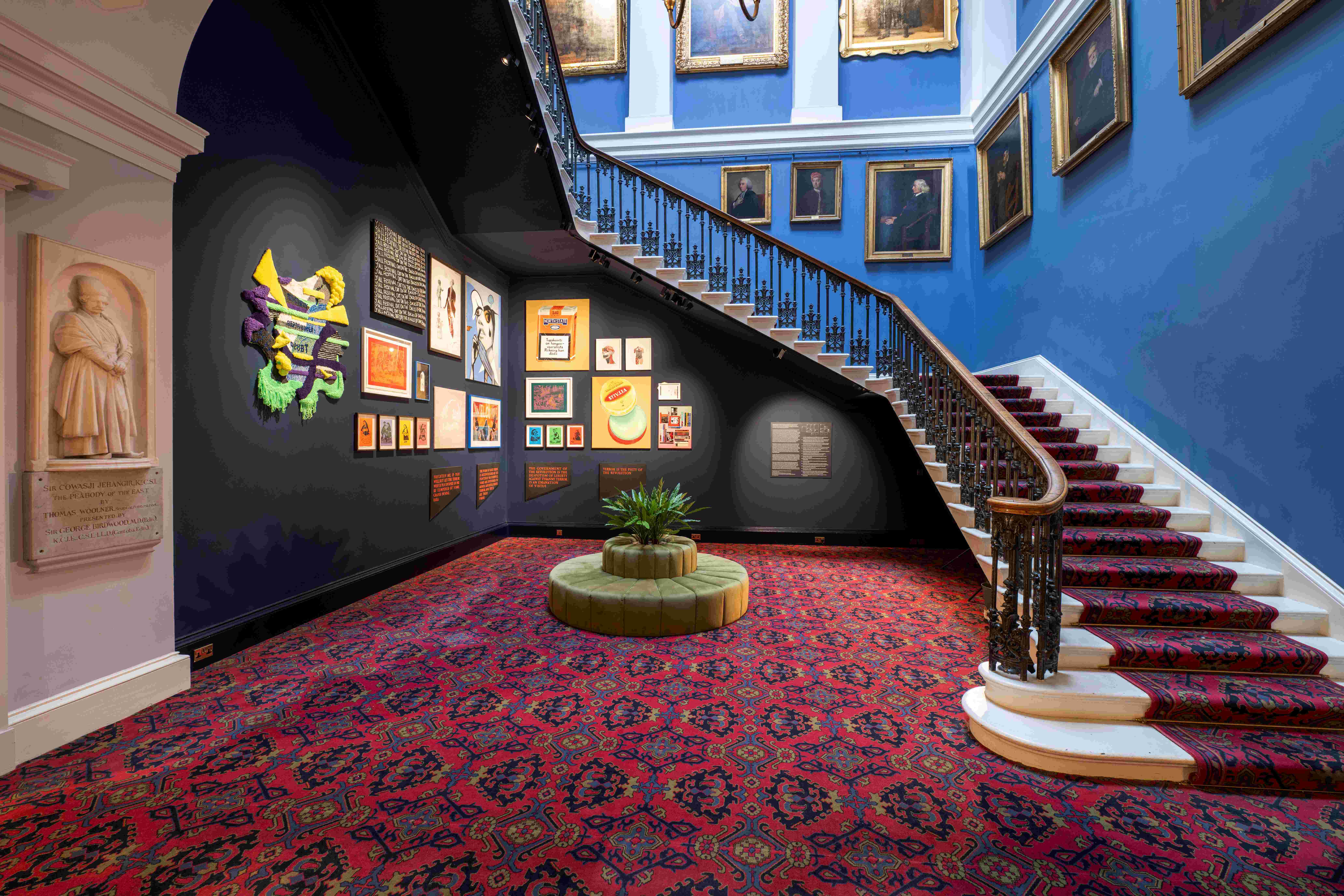 Display of art works at the Old College of the University of Edinburgh. The display is located below a staircase and consists of a variety of vibrant and colourful pieces. There is a plant across the artwork. We can see the portraits of thinkers and professors and principals in the background.