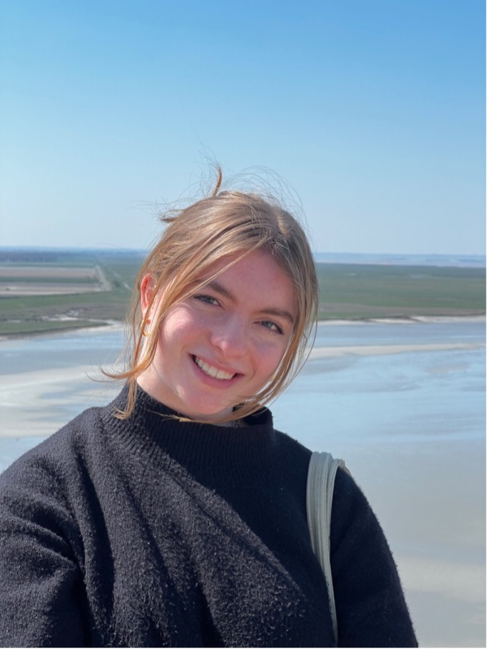 young woman with blonde hair smiles into the camera, a sea behind
