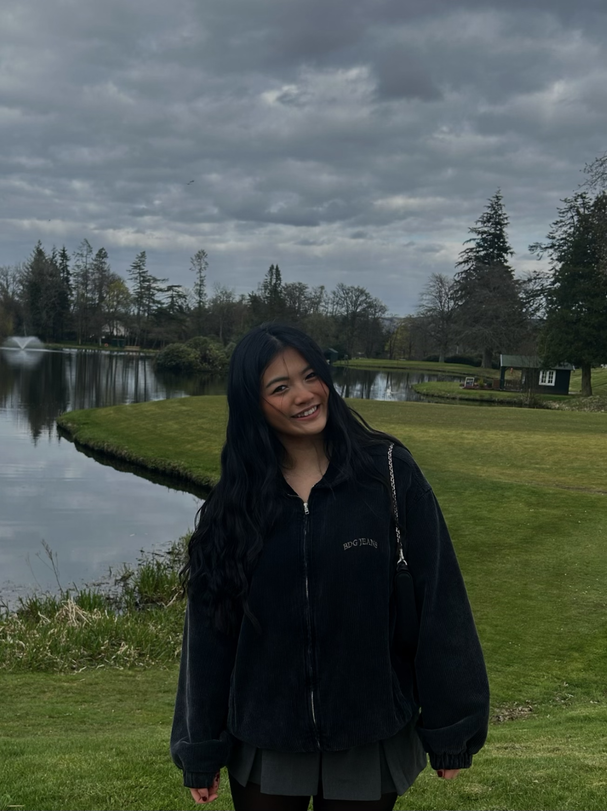 A Picture of Maggie Outside by a Lake with Trees in the back ground. She wears a black zip-up jumper