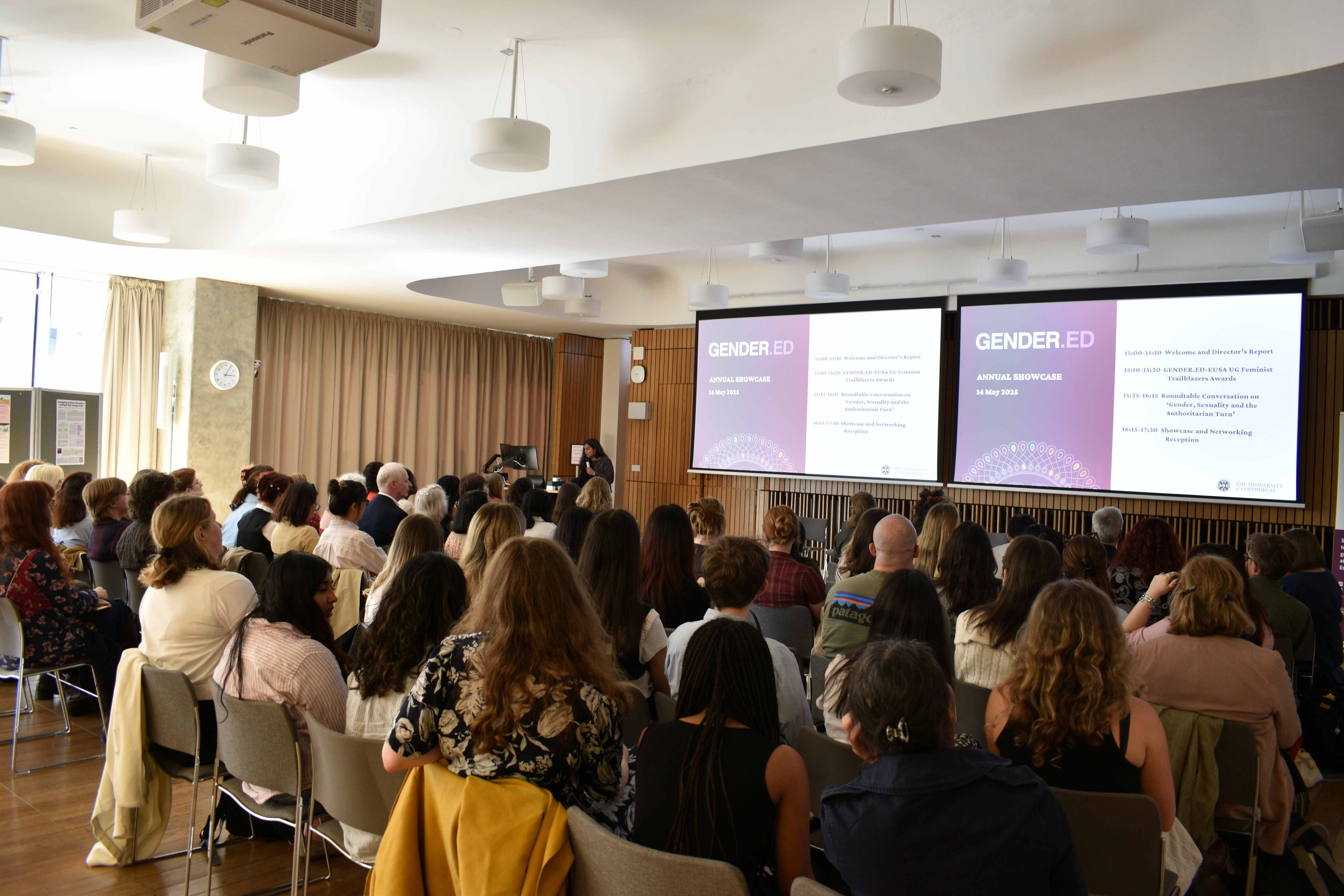 Photo of Radhika Govinda, director of GENDER.ED, speaking in front of rows of people at the Annual Research Showcase. She is holding a microphone. To her right, there is a screen that with a slide that reads 'GENDER.ED'.