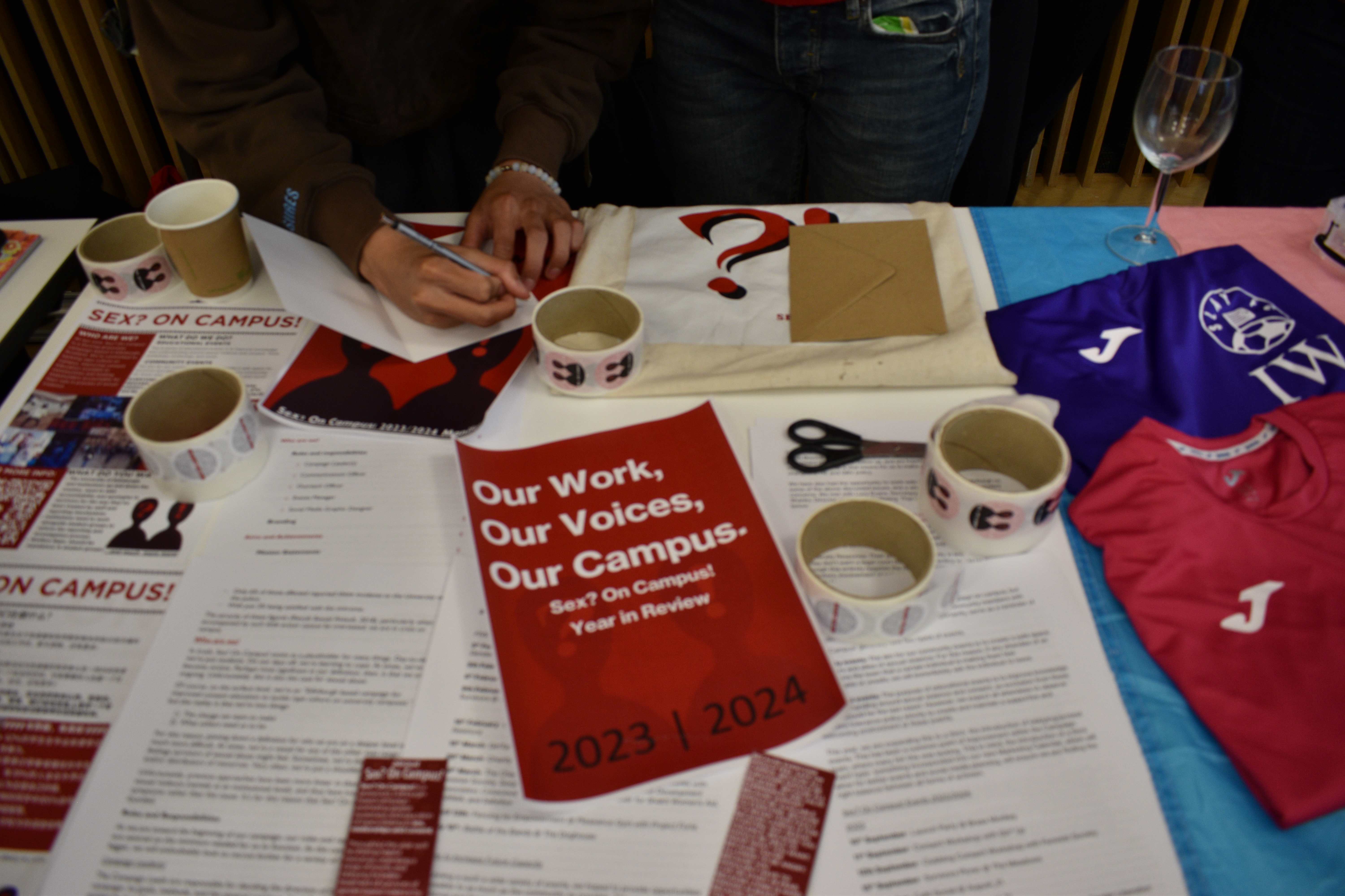 Sex? On Campus! table at the Annual Research Showcase. A lot of papers, stickers, and documents are displayed, with one of them reading 'Our Work, Our Voices, Our Campus'. At the back, one of the members of the team is writing on a piece of paper.