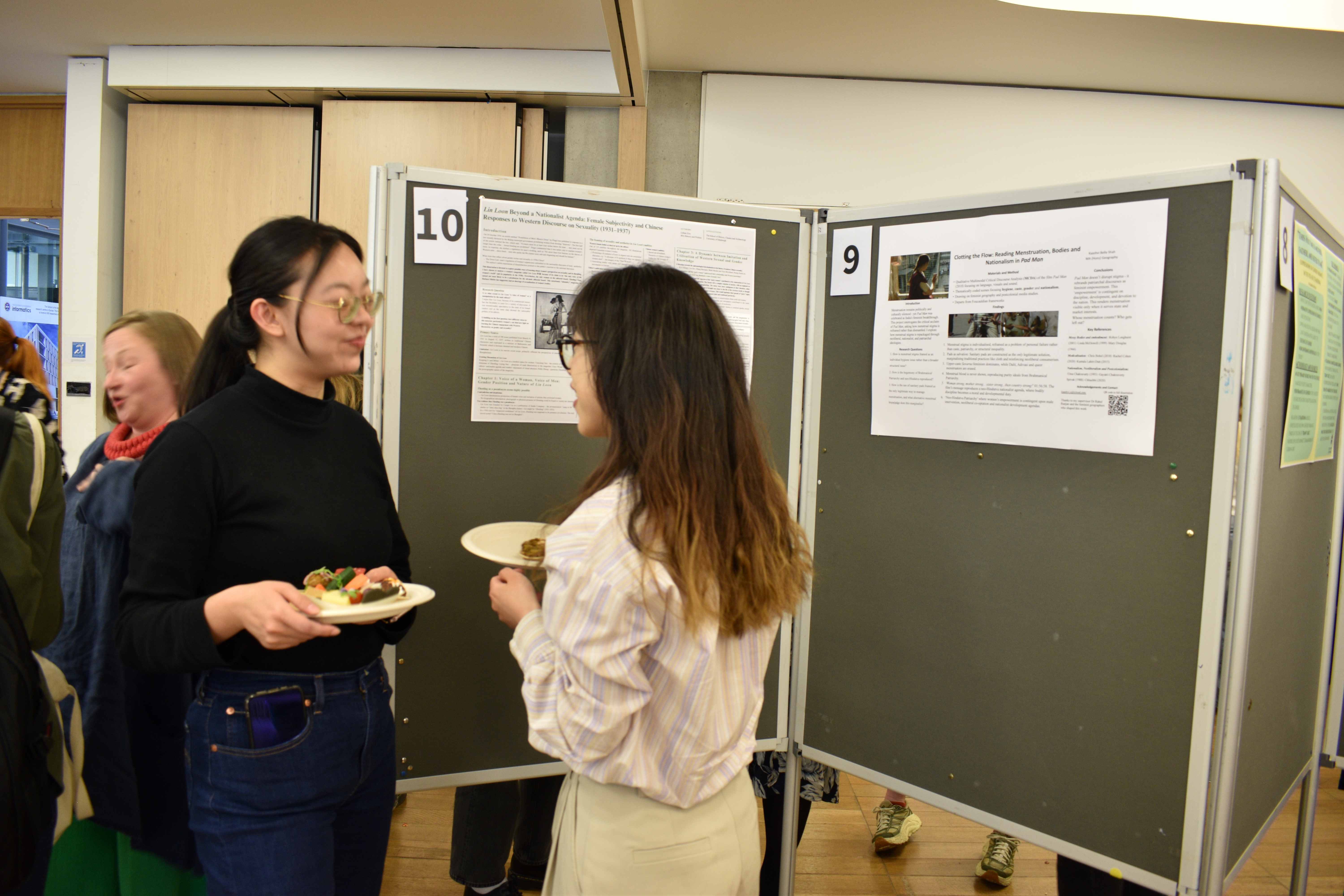 Photo of two undergraduate students chatting and having a bite in front of their posters at the mini undergraduate dissertation showcase.