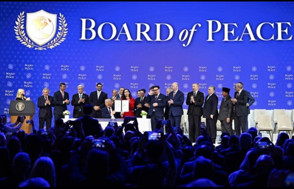 Donald Trump holding up the Board of Peace Charter amid a crowd of board members on stage in front of a media crowd at the Announcement and Signing Ceremony in January 2026